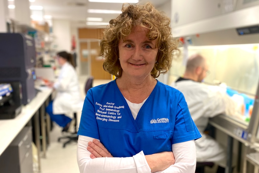 A woman stands in a hospital in blue scrubs and a white undershirt.